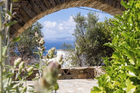 Un arco de piedra que da a vistas del mar, con olivos y un muro de piedra al frente.