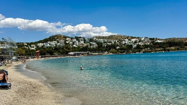 Une plage de galets bordée d’une eau cristalline, une ville lointaine et un ciel dégagé.