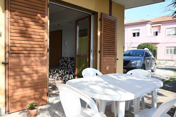 Une porte en bois ouverte donne sur un patio abrité, où se trouvent une table et des chaises blanches, une voiture garée et, au fond, un bâtiment rose.