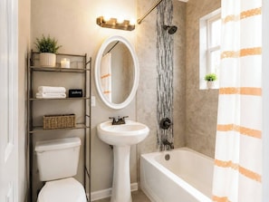 A bathroom with a toilet, pedestal sink, bathtub, and a shelf with towels and a makeup case.