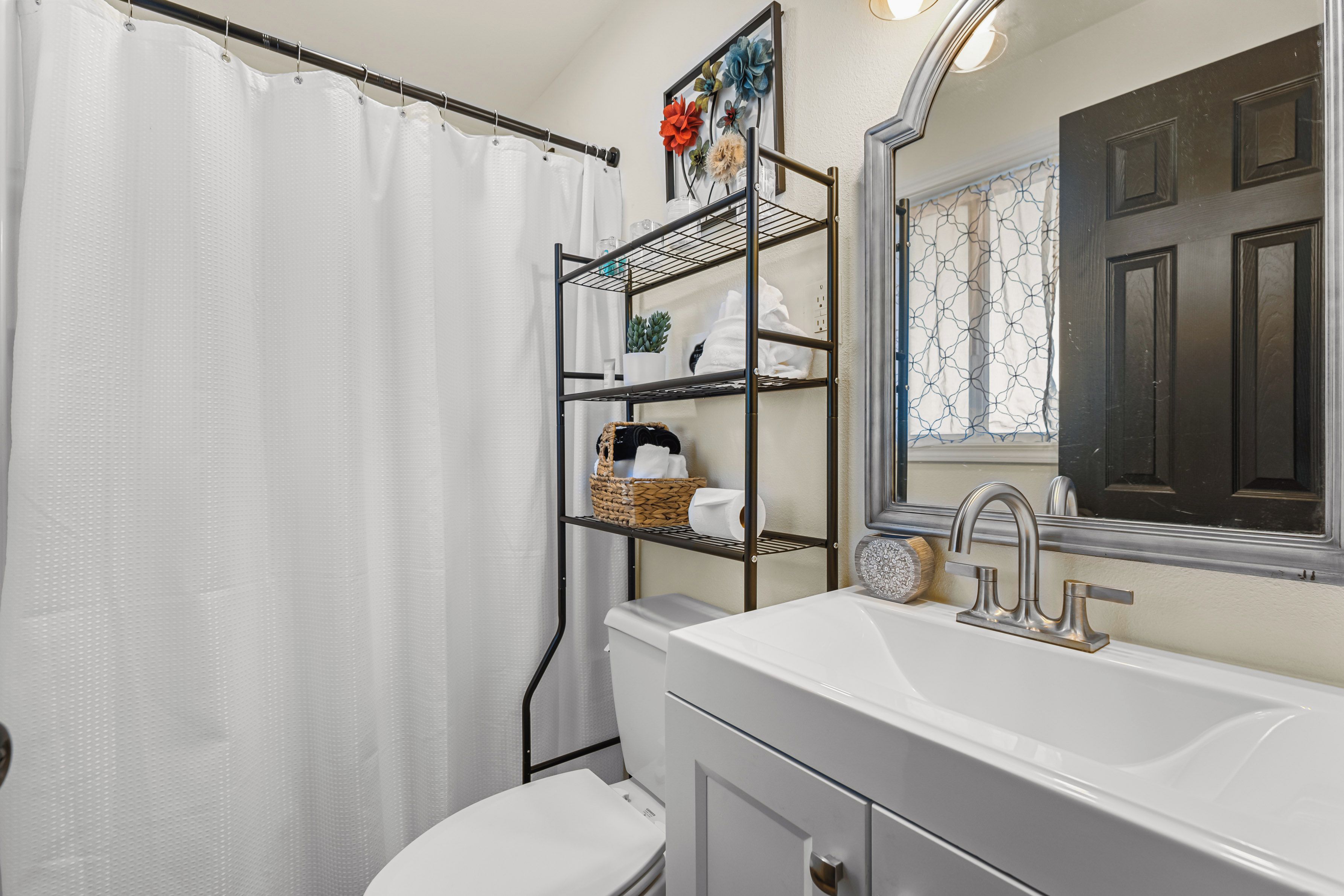 A bathroom with a white toilet, a white sink with a modern faucet, a metal shelf unit with baskets, and a mirror above the sink.
