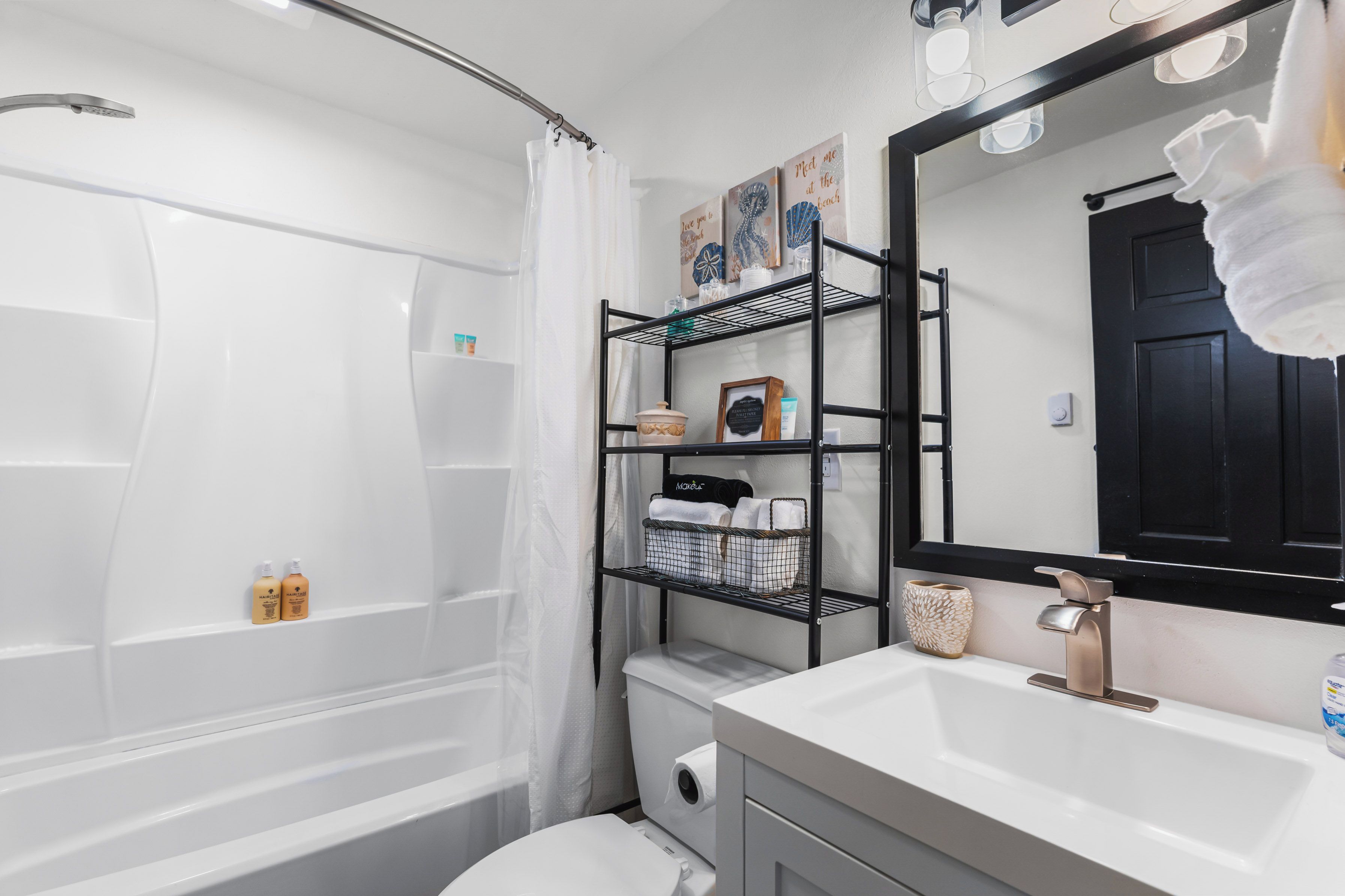 A modern bathroom with a white bathtub, a vanity with a sink, and a black shelving unit above the toilet.