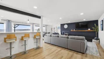 A modern living room with a grey sofa, wooden bar stools, a fireplace, and a large window.