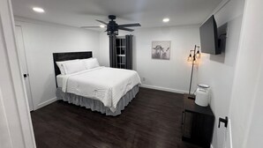 Une chambre à coucher avec un lit, un ventilateur de plafond, un téléviseur fixé au mur et une fenêtre avec des rideaux.