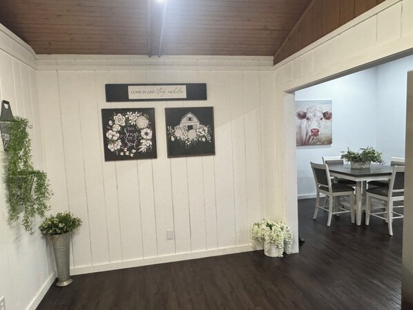 A dining area with a wooden ceiling, white walls, and a dark wooden floor. There are two framed artworks on the wall, a potted plant, and a dining table with chairs.