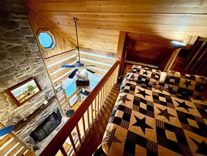 A room with a wooden ceiling, a bed with a star-patterned quilt, a stone wall, and a ceiling fan.