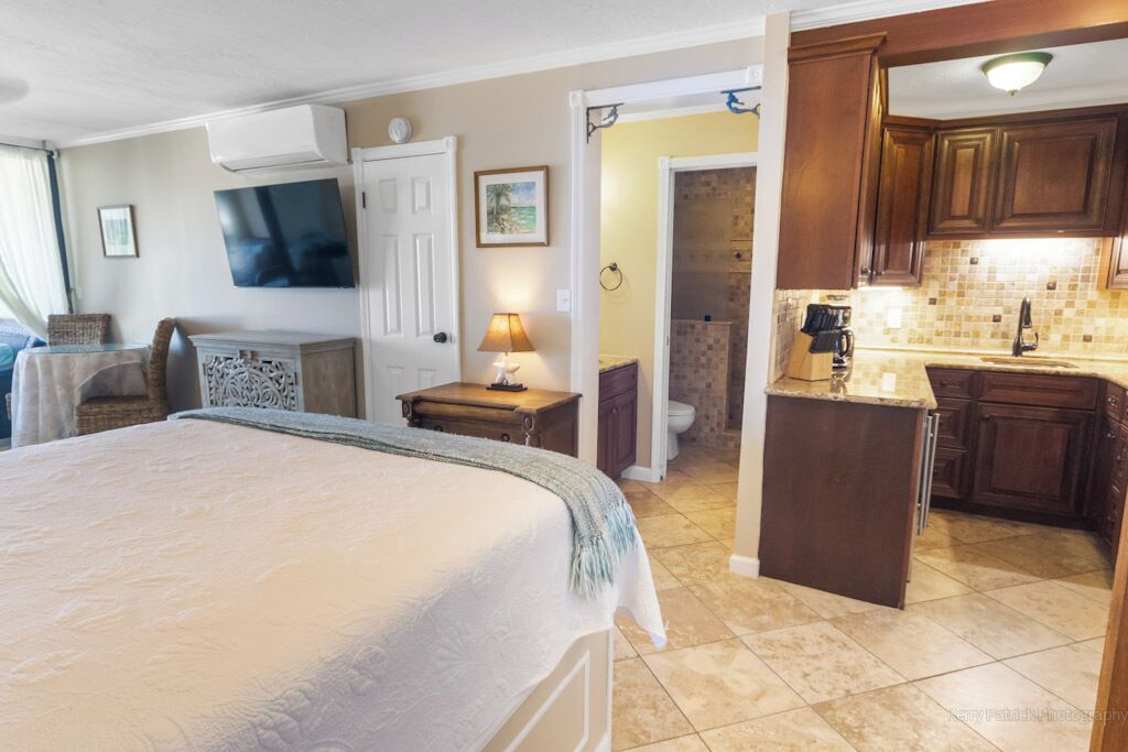 A bedroom with a large bed, a bedside table with a lamp, a television mounted on the wall, and a view into the kitchen area with wooden cabinets and a tiled backsplash.