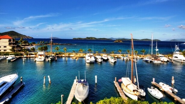Un port de plaisance où sont amarrés de nombreux bateaux, un ciel bleu azur et une vue lointaine sur un paysage montagneux.
