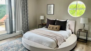 Ein rundes Bett in Beige- und Weißtönen, zwei Nachttische mit Lampen, ein großes Fenster mit Blick ins Grüne und ein gerahmtes Bild an der Wand.