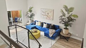 Ein modernes Wohnzimmer mit einem blauen Modulsessel, einem gelben Hocker und zwei großen Kübelpflanzen.