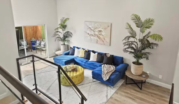 Un salon moderne avec un canapé d’angle bleu, un pouf jaune et deux grandes plantes en pot.