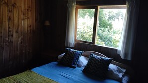 Una stanza con parete in legno, un letto con biancheria azzurra e cuscini decorati, una finestra con tende bianche e vista sul verde circostante.