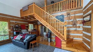Ein Ferienhaus-Interieur mit einer Holztreppe, einem Wohnbereich mit einer Couch, einer Küche und einem großen Fenster mit Blick auf Grünflächen.