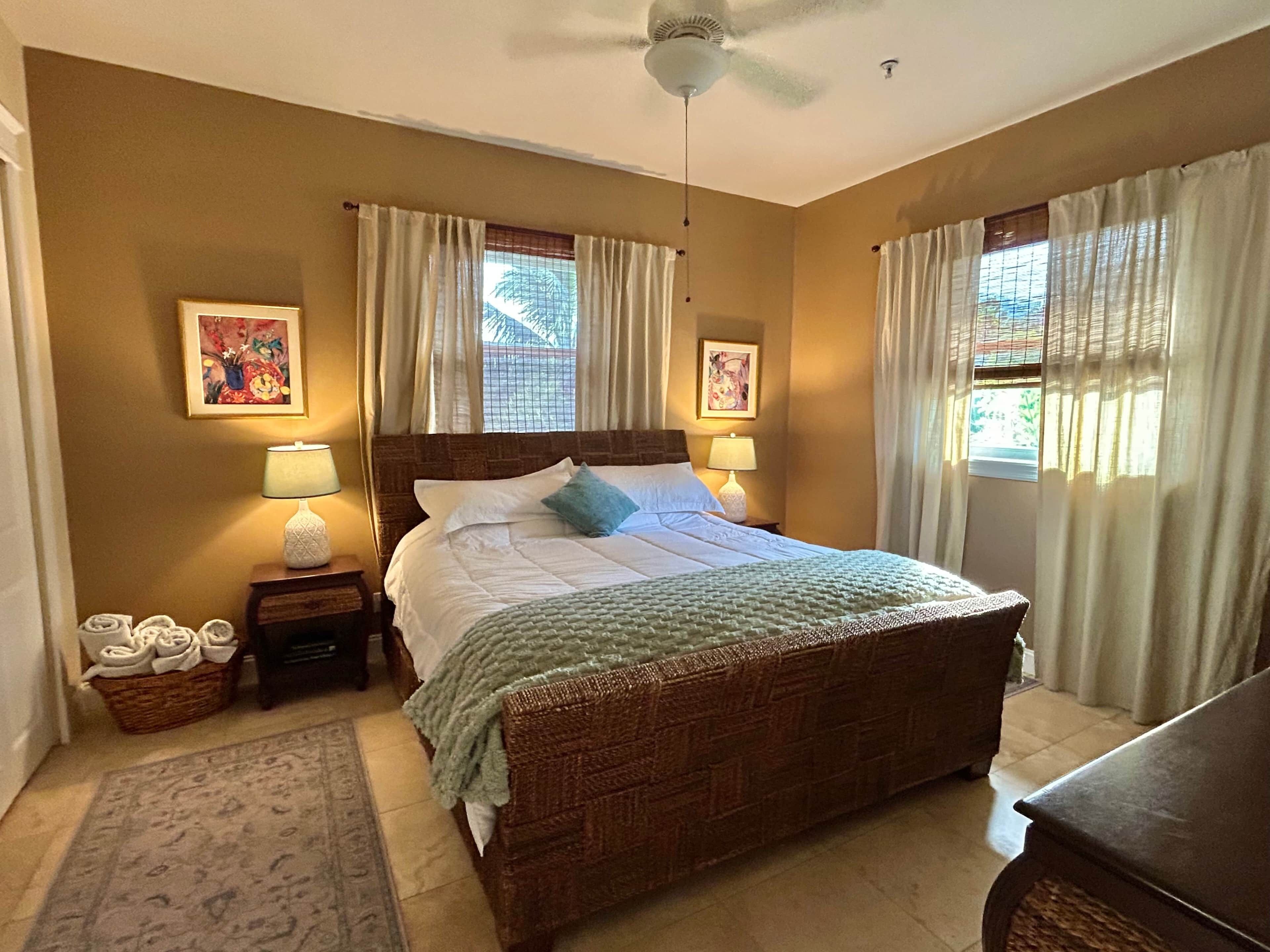 A bedroom with a large bed, two bedside lamps, a ceiling fan, and two windows with curtains.