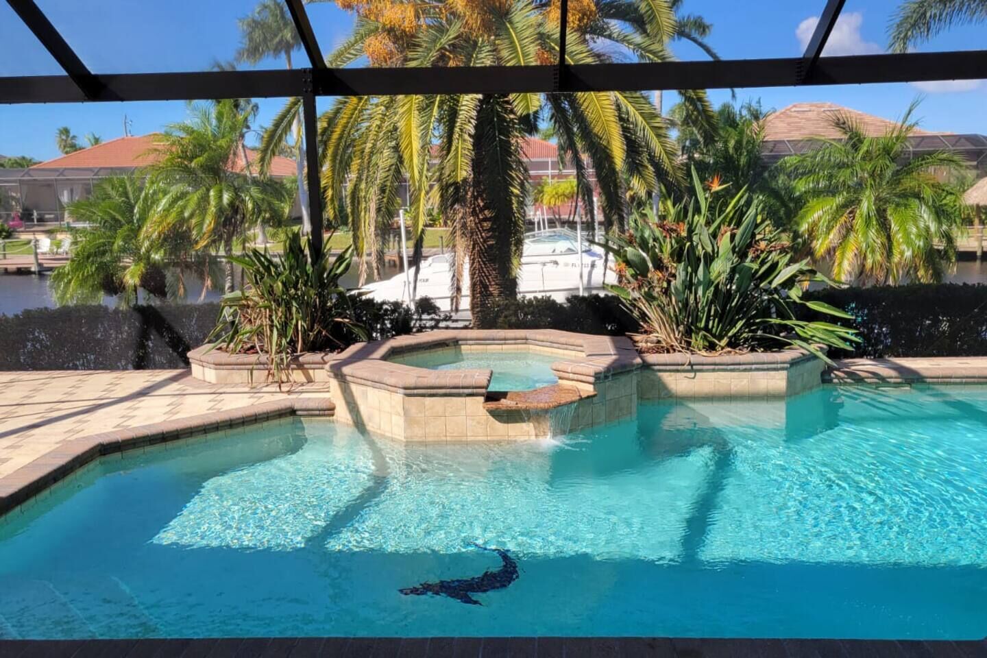 A swimming pool with a hot tub, surrounded by palm trees and a view of a building with a red roof.