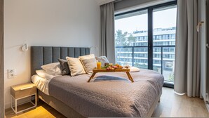 Une chambre d’hôtel moderne avec un grand lit, une table de chevet, un plateau avec des articles pour le petit-déjeuner et une vue sur la ville grâce à de grandes fenêtres.