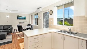 A modern kitchen with a large window, a dining area with a table and chairs, and a living room with a sofa and a TV.