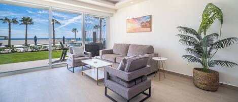 A modern living room with a grey sofa, a small table, and a chair. There is a large window with a view of palm trees and a pool. A painting hangs on the wall, and a large plant is in a woven basket.