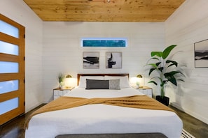A bedroom with a wooden ceiling, a large bed with white bedding, two bedside tables with lamps, a window with a view of trees, and two abstract paintings on the wall.