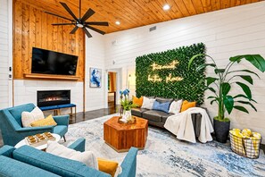 A modern living room with a fireplace, a flat-screen TV, a ceiling fan, and a green wall with a 'Christmas is the place' sign. - 1-Story Stay | Hot Tub, PingPong, Arcade & Playset (Broken Bow)