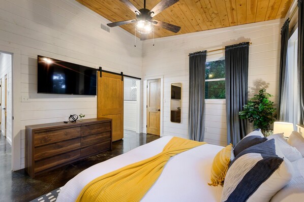 A bedroom with a bed, a wooden dresser, a ceiling fan, a TV mounted on the wall, and a window with curtains.