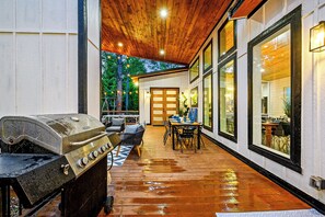 Een terras buiten met een barbecue, een eethoek met een tafel en stoelen, en een uitzicht op het interieur door grote ramen.