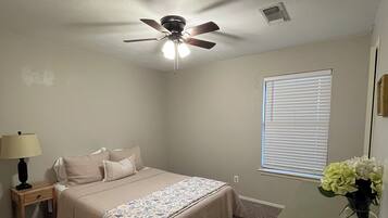 A bedroom with a bed, a nightstand, a lamp, a ceiling fan, a window with blinds, and a vase with flowers.