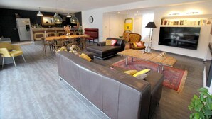 A modern living room with a sectional sofa, a dining area with chairs, a kitchen in the background, and a flat-screen TV mounted on the wall. - Near Uzès, holiday air-conditioned villa located Vers-Pont-du-Gard, private pool. (VERS-PONT-DU-GARD)
