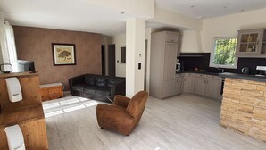 Un salón amplio con un sofá de cuero oscuro, un sillón marrón y una mesa de centro de madera. Se observa una cocina con isla de piedra y armarios al fondo.