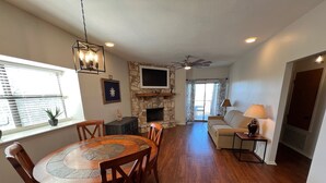 A spacious living room with a fireplace, a dining area with a wooden table and chairs, a sofa, and a TV mounted on the wall. - Peaceful river views/Guadalupe access/Swimming Pool/2 bed 2 bath/Sleeps 6 (New Braunfels)