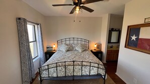 A bedroom with a bed, a ceiling fan, a window with curtains, a nightstand with a lamp, and a framed picture on the wall. - Peaceful river views/Guadalupe access/Swimming Pool/2 bed 2 bath/Sleeps 6 (New Braunfels)