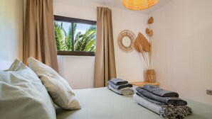 A room with a bed, curtains, a window with a view of palm trees, a woven wall decoration, and folded towels. - Oasis Duna-Pistacho Dreams Pool: Shared Pool, Private Terrace & Wi-Fi (corralejo)