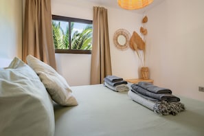 Una habitación con cama, cortinas, una ventana que da a palmeras, un adorno de pared tejido y toallas dobladas.