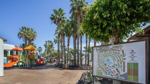 A playground with slides and climbing structures, palm trees, and a map sign. - Oasis Duna-Pistacho Dreams Pool: Shared Pool, Private Terrace & Wi-Fi (corralejo)