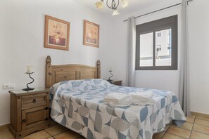 Une chambre à coucher avec un lit en bois, une table de chevet, une lampe de chevet, deux lampes fixées au mur et une fenêtre donnant sur les bâtiments environnants.