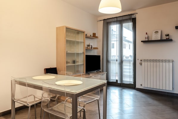 A modern dining area with a glass table, clear chairs, a wooden cabinet, a television, and a sliding glass door leading to a balcony.