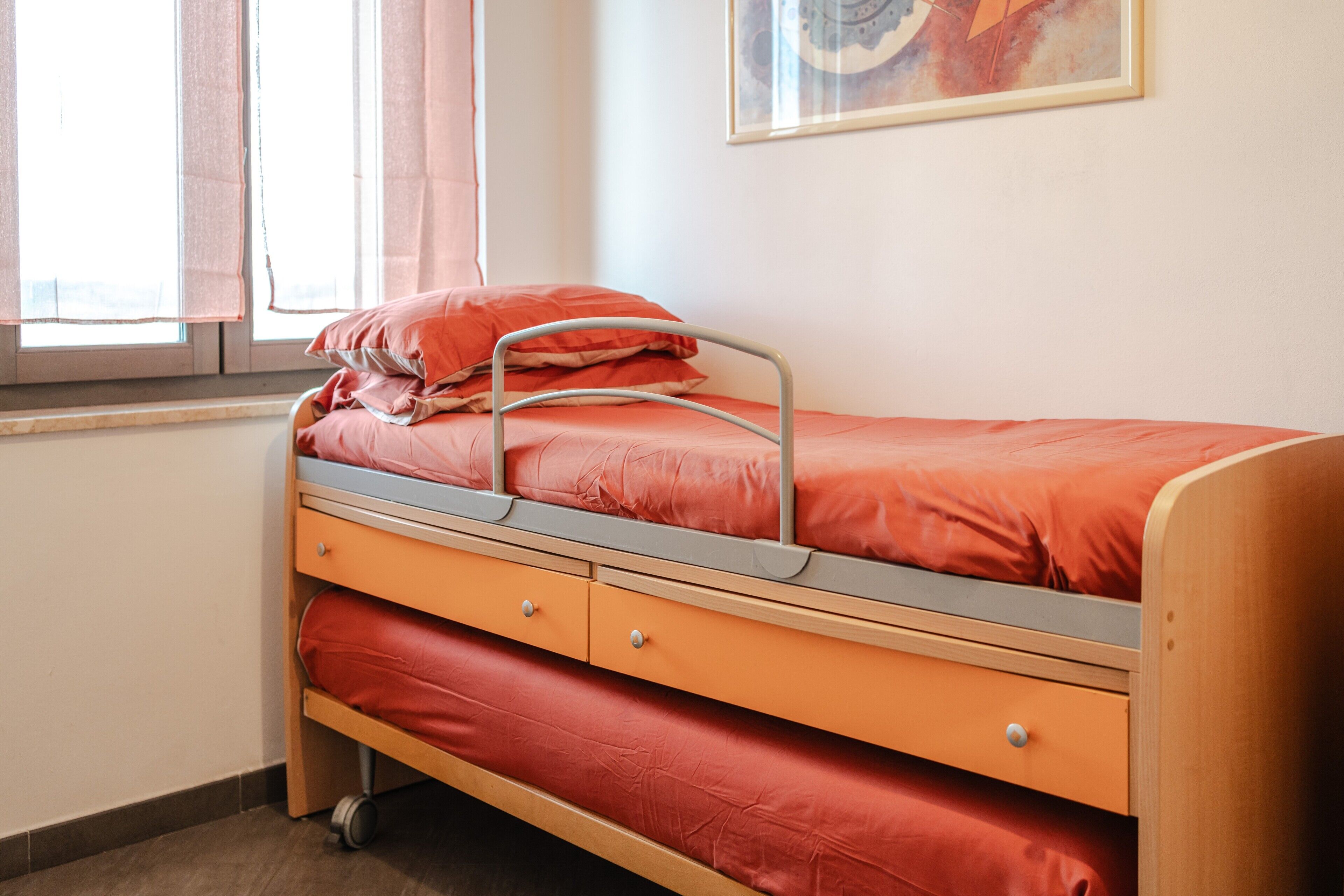 Una cama individual con estructura metálica, cabecera de madera y colchón rojo. Se aprecia una ventana con cortinas, una obra de arte en la pared y un cajón pequeño debajo de la cama.