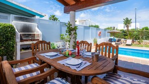 Un espace extérieur aménagé pour les repas, avec une table et des chaises en bois, un barbecue et une piscine en arrière-plan.