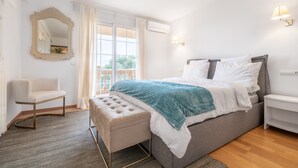 Un dormitorio con cama, una silla, un banco, un espejo y una ventana con cortinas.