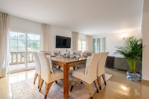 Un coin repas avec une table en bois et des chaises en osier, un téléviseur à écran plat fixé au mur et une grande plante en pot.