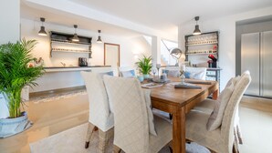 Un comedor moderno con una mesa de madera, sillas acolchadas, iluminación colgante y una cocina al fondo.