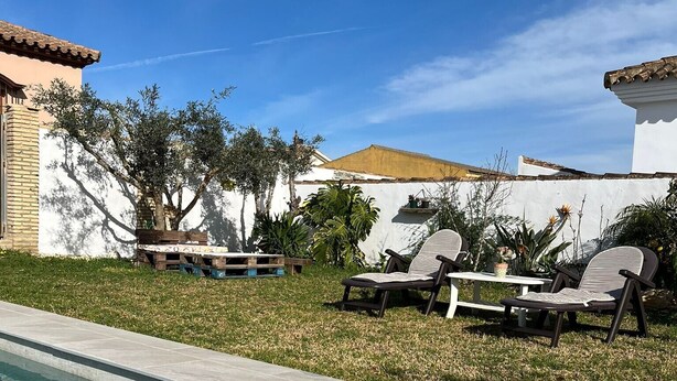 Una piscina con borde de azulejos, una zona verde y un edificio blanco con techo de tejas.