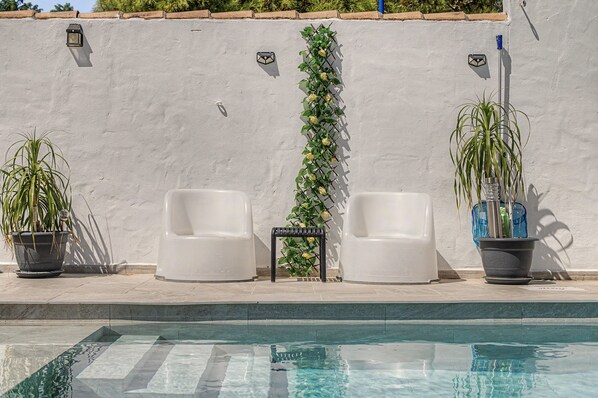 Un área junto a la piscina con dos sillones blancos, una mesita y plantas en macetas.