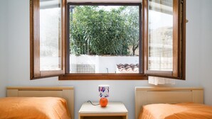 A room with two beds, a small table with a mug, and a window with a view of greenery.