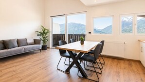 En moderne stue med trægulv, en grå sofa, et spisebord med stole og store vinduer med udsigt til bjerge.