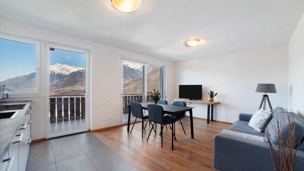 En moderne stue med sofa, spisebord, stole, et fjernsyn og en balkon med udsigt til bjerge.