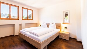 A bedroom with a wooden bed, a nightstand, a bedside lamp, and three windows with a mountain view. - Apartment 'Preiss Type B' with Mountain View, Balcony and Wi-Fi (Schenna)