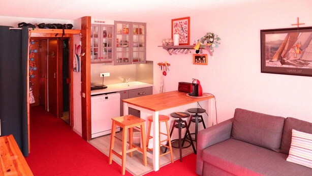 Een compacte leefruimte met een kitchenette, een eethoek met krukken, een bank en een rood tapijt.