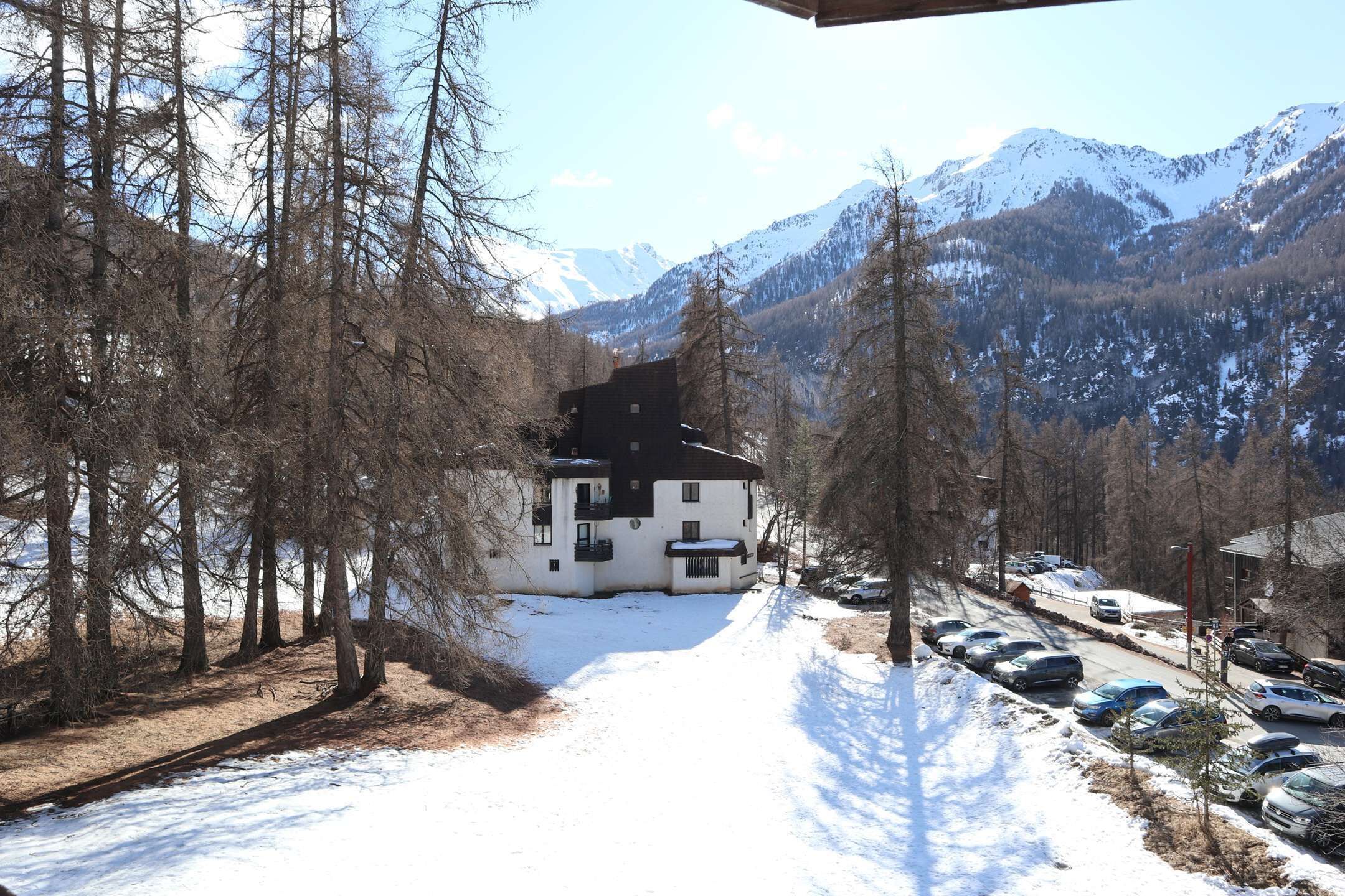 Une route enneigée mène à un bâtiment blanc, surmonté d’un toit noir, et qui est entouré d’arbres. On aperçoit des montagnes au loin.