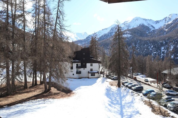A snow-covered road leading to a white building with a black roof, surrounded by trees and mountains in the background.